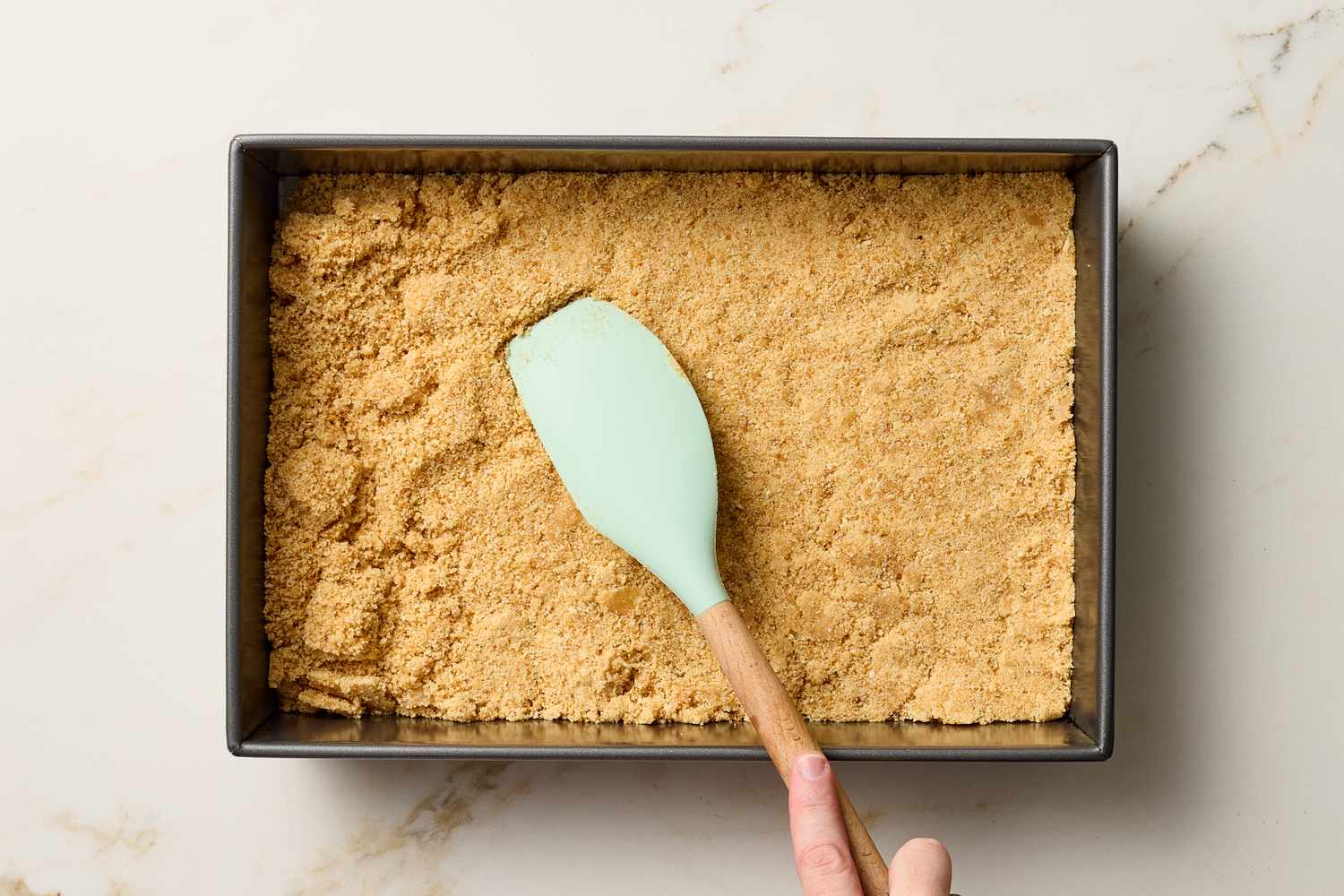 Crumbly crust mixture being pressed into a rectangular pan with a spatula
