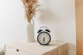 An alarm clock on a wooden surface next to a vase with dried plants