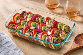 Dish of sliced vegetables arranged in rows in a baking dish with garlic cloves scattered on top