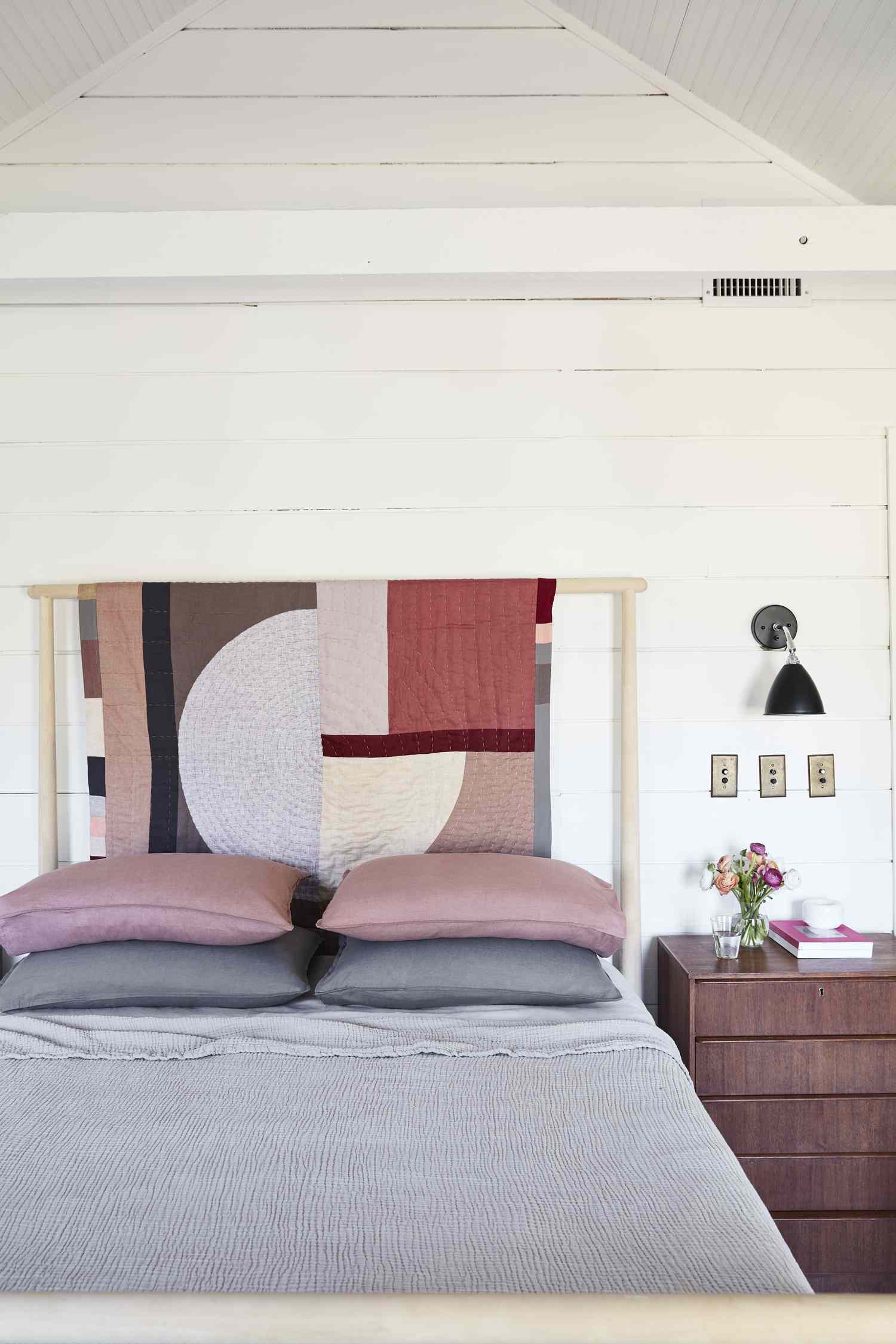A modern bedroom with a bed featuring a colorful quilted wall hanging as a headboard backdrop and a nightstand with decor