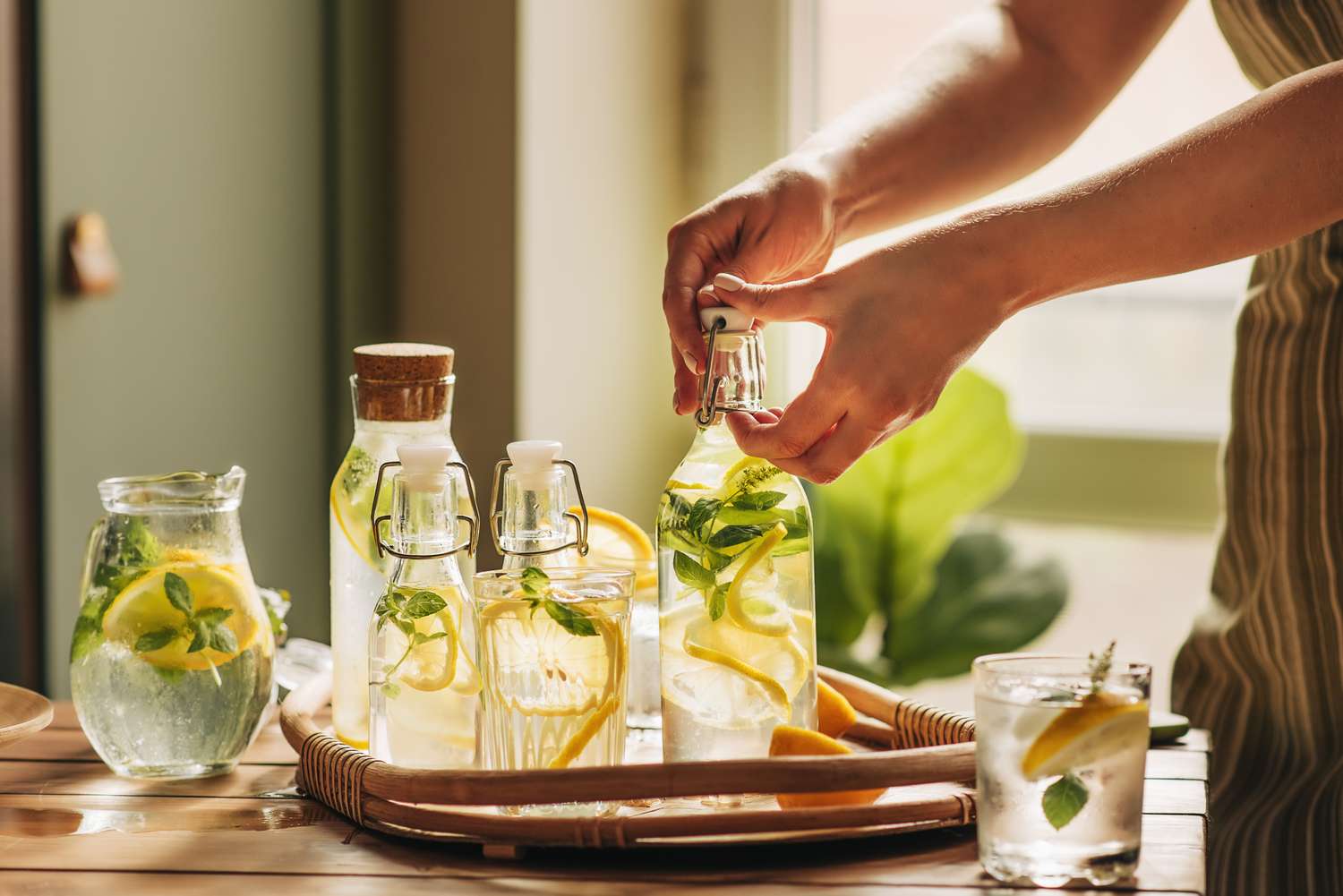 Person preparing bottles and glasses with lemon slices and herbs on a tray