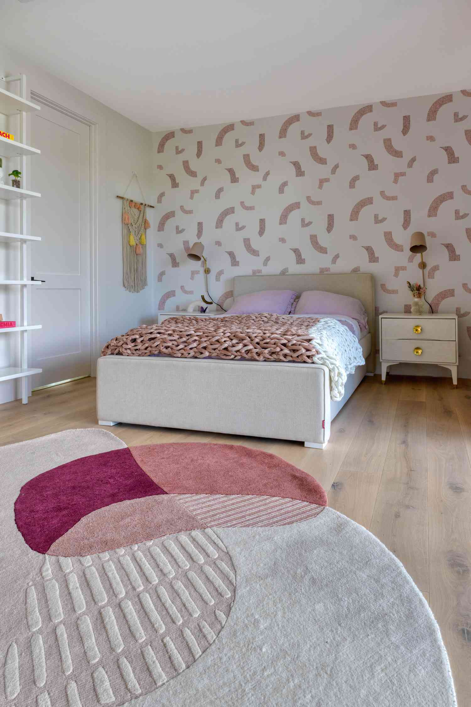 A queen bedroom with a pink and cream color scheme. Overlapping kidney-shapes in the rug echo similar forms on the white and pink wallpaper.