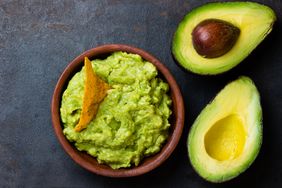 A bowl of guacamole with a tortilla chip accompanied by two avocado halves on a dark background