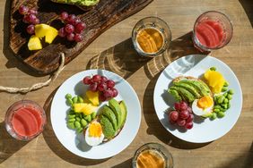 Two plates with avocado halfboiled egg grapes pineapple edamame and served with glasses of juice and coffee on a table