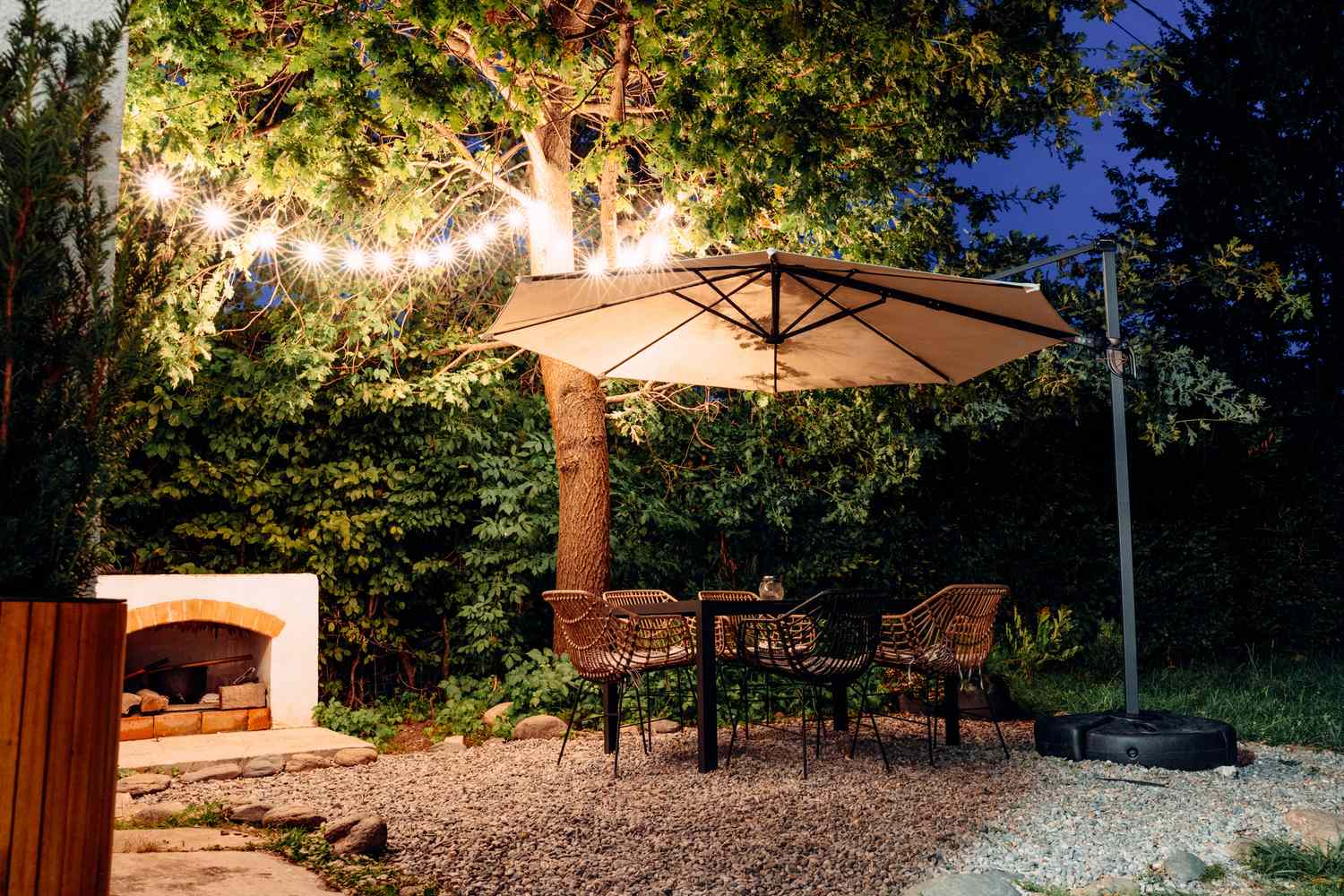 Outdoor patio with chairs around a table under an umbrella, near a tree and illuminated string lights