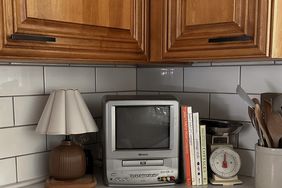 A small television sits on a kitchen counter surrounded by various items including books a lamp and a kitchen scale