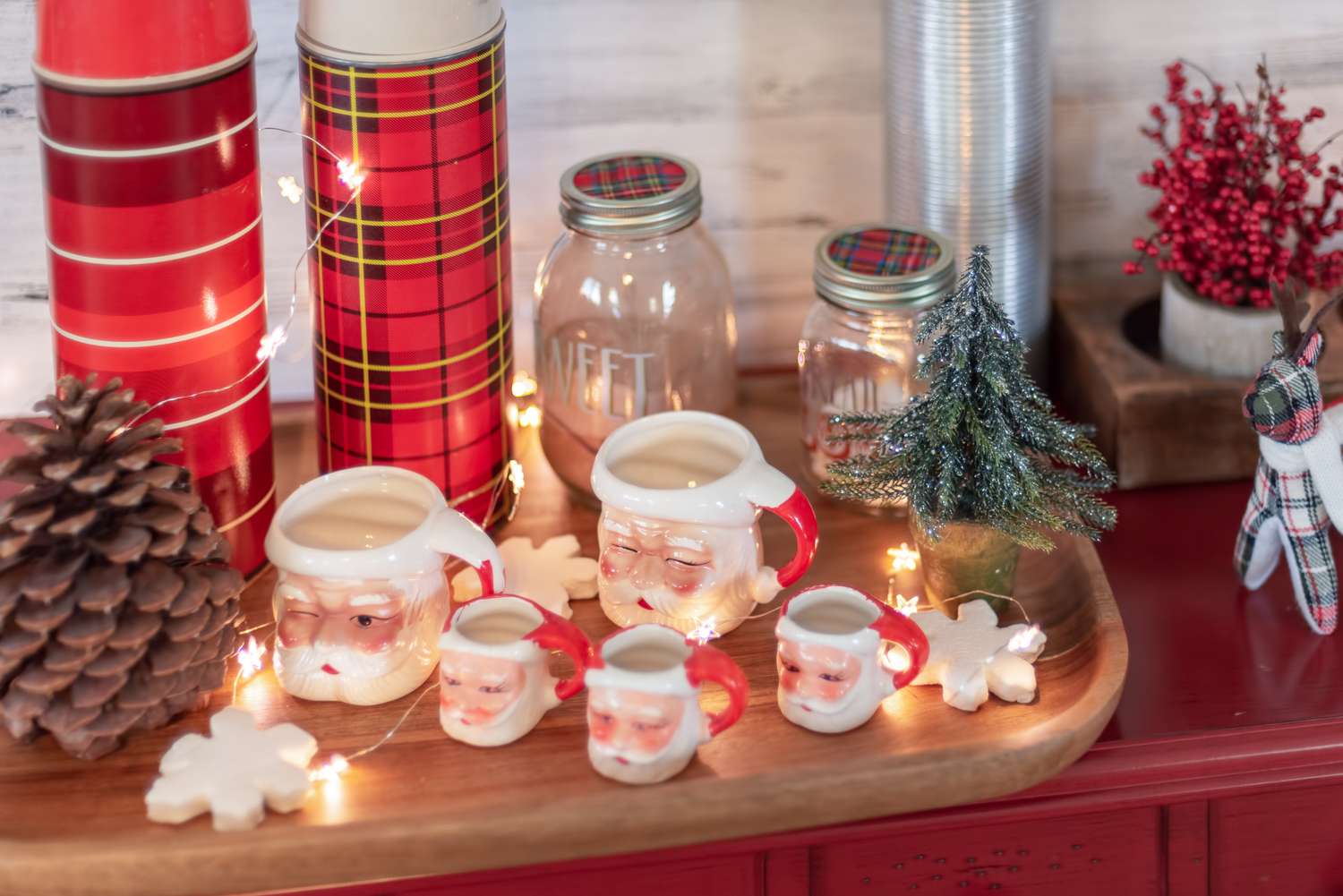 A display of Santathemed mugs pinecones mason jars and Christmas decor such as a small tree and star lights on a wooden tray