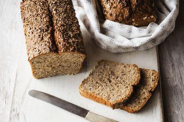 loaf of seeded bread and some slices on cutting board with knife, and a loaf of oat bread in a cloth