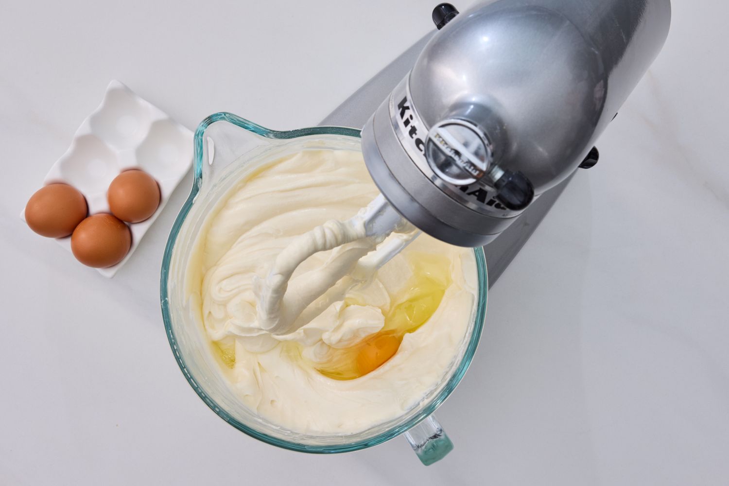 A stand mixer with a glass bowl mixing cheesecake batter and an egg three whole eggs in a carton are beside it