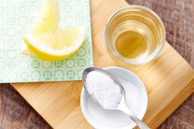 A small bowl of white powder with a spoon a glass of liquid and lemon wedges on a wooden board