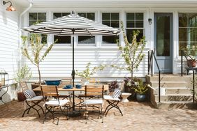 Outdoor patio area with a table chairs and an umbrella against a white house with lush plants and windows in the background