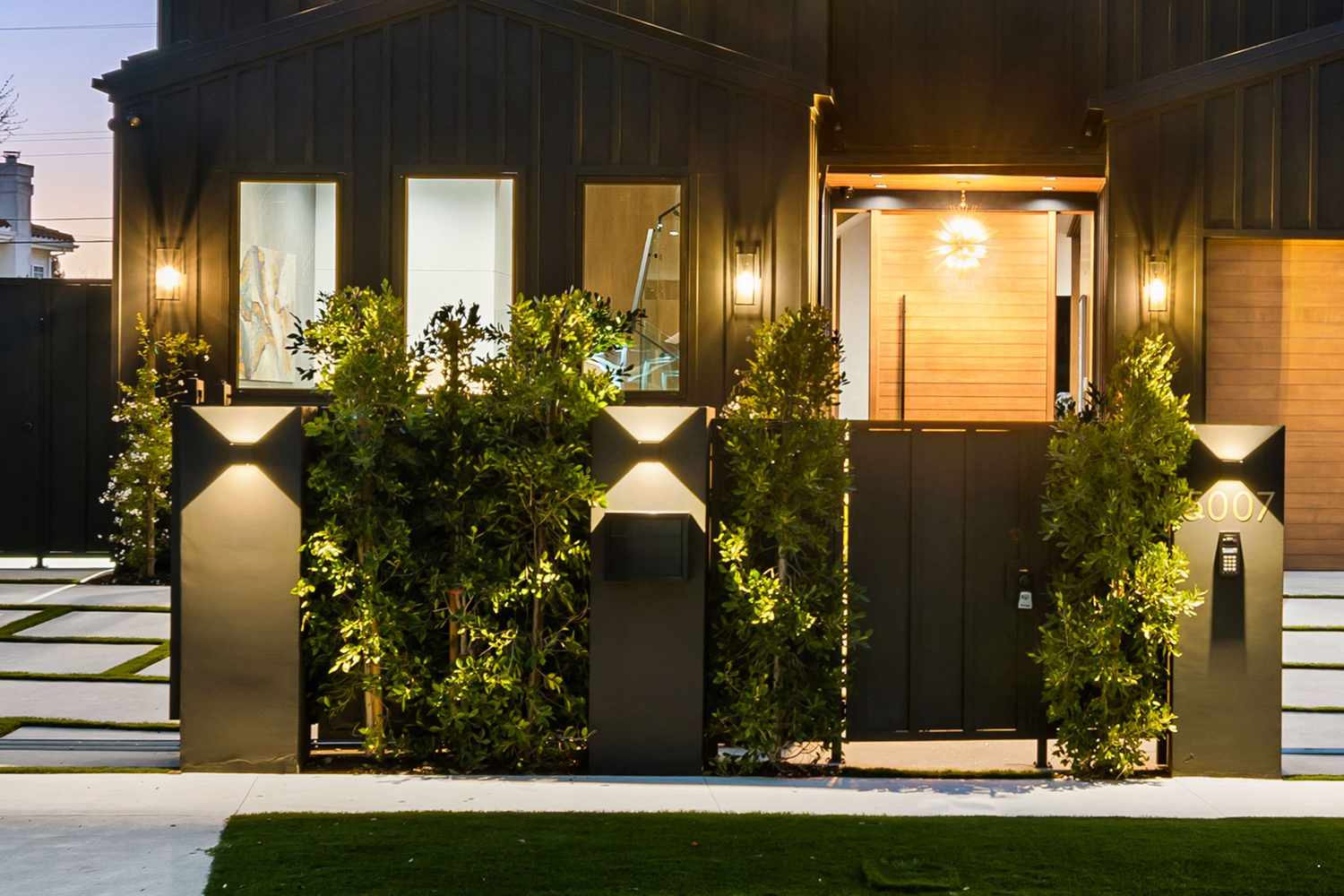 A contemporary residential entryway with modern lighting and landscaping featuring a dark exterior and illuminated greenery