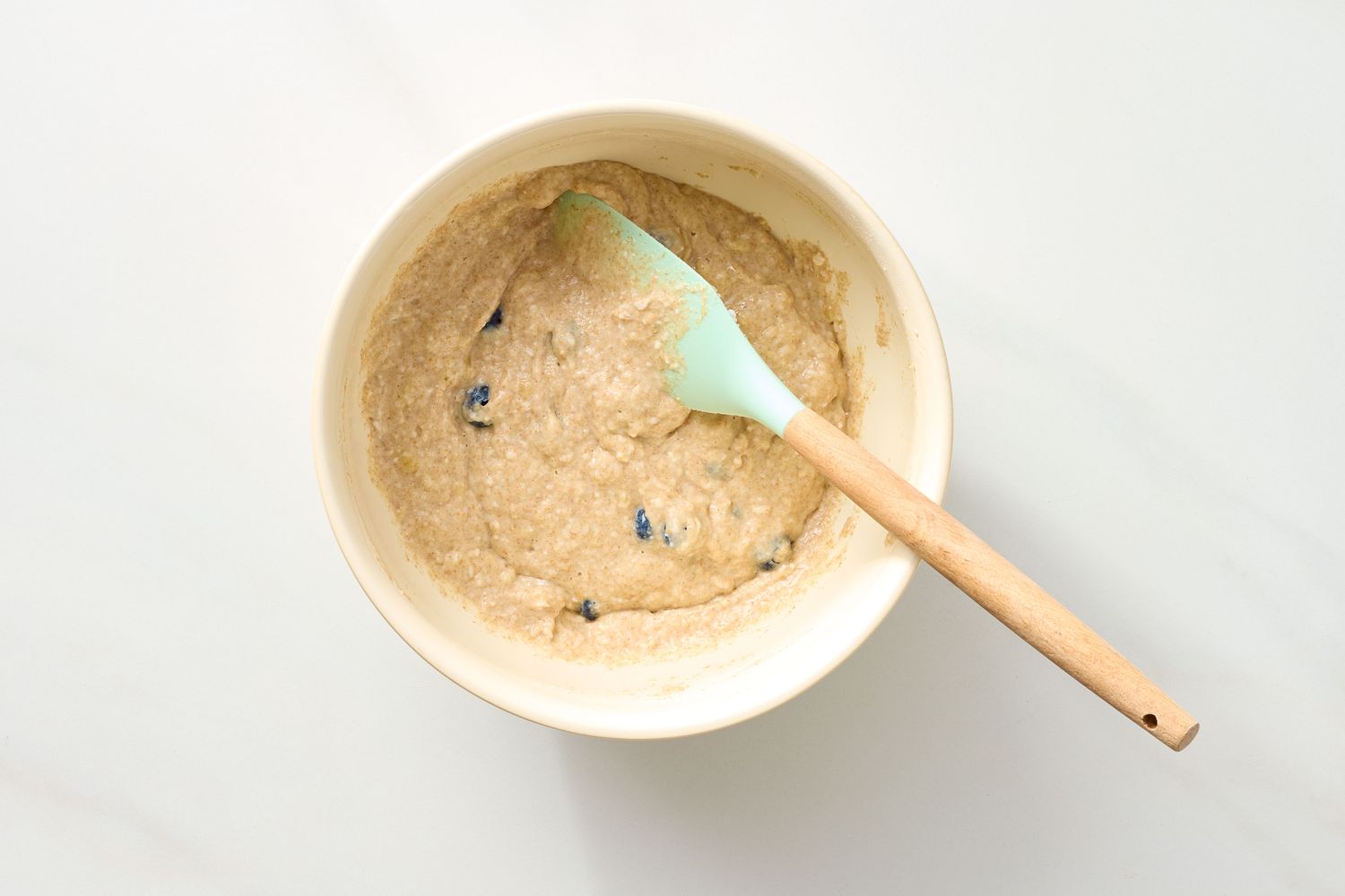 Bowl with muffin batter mixed with blueberries and a spatula