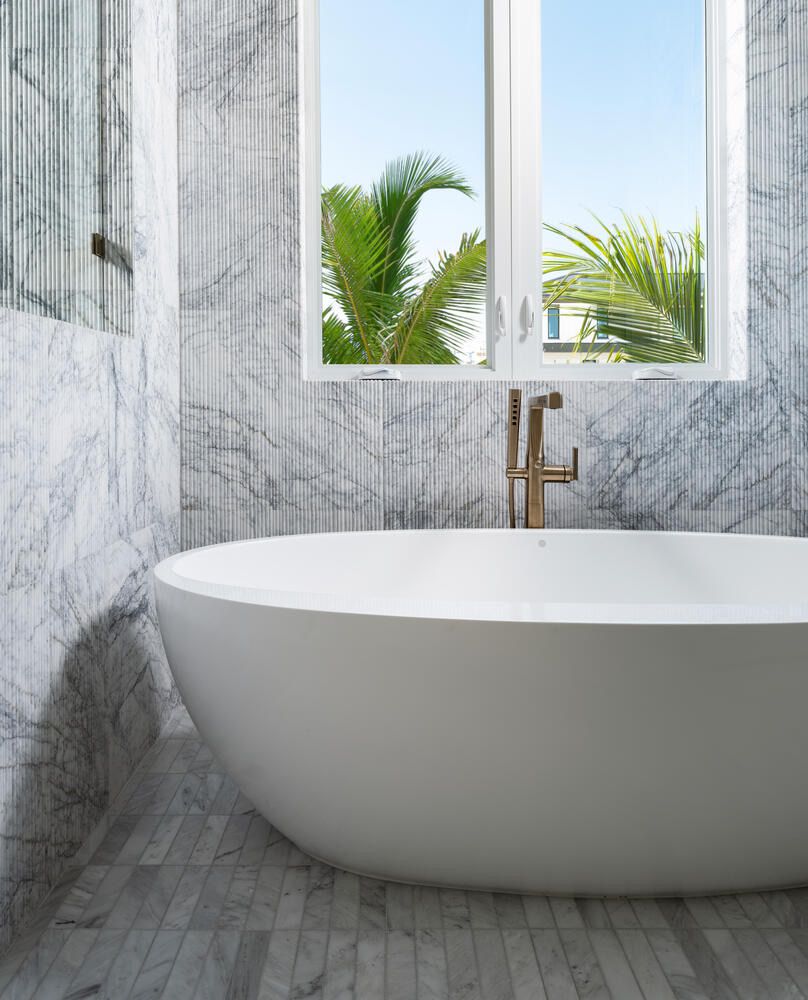 Freestanding bathtub in a marble-lined bathroom with a large window showing palm trees outside