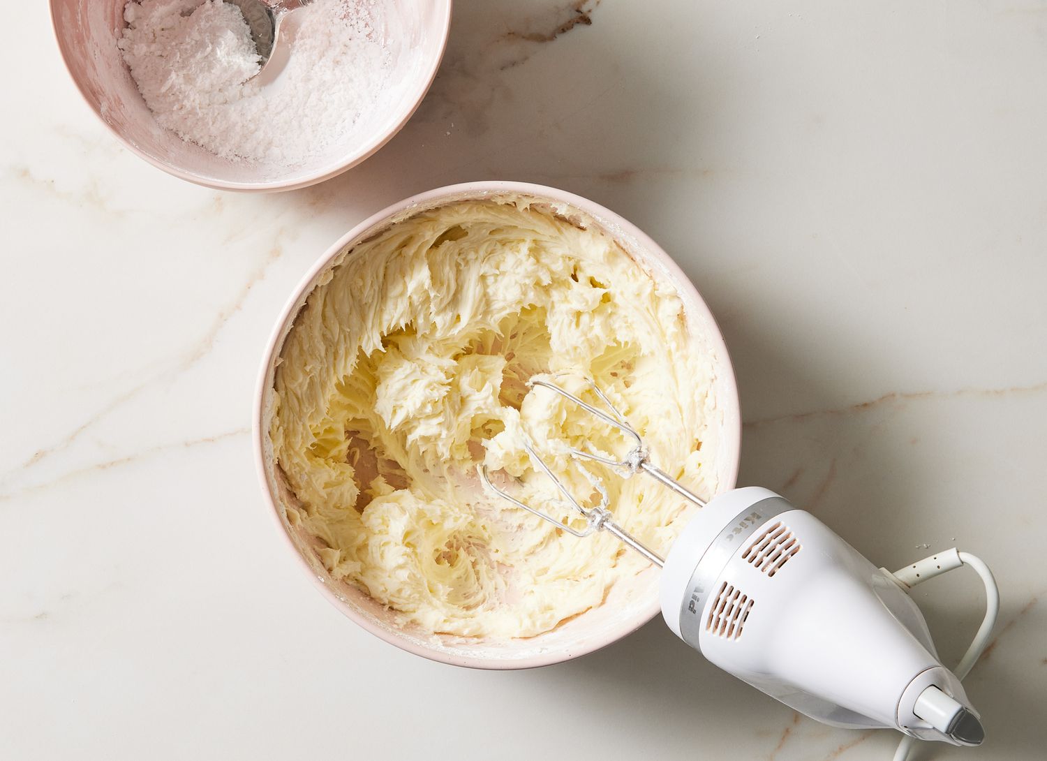 overhead view of mixing bowl of white frosting, hand mixer and bowl of flour