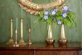 A wooden table with brass vases holding flowers and candlesticks a framed mirror on a greenpaneled wall in the background