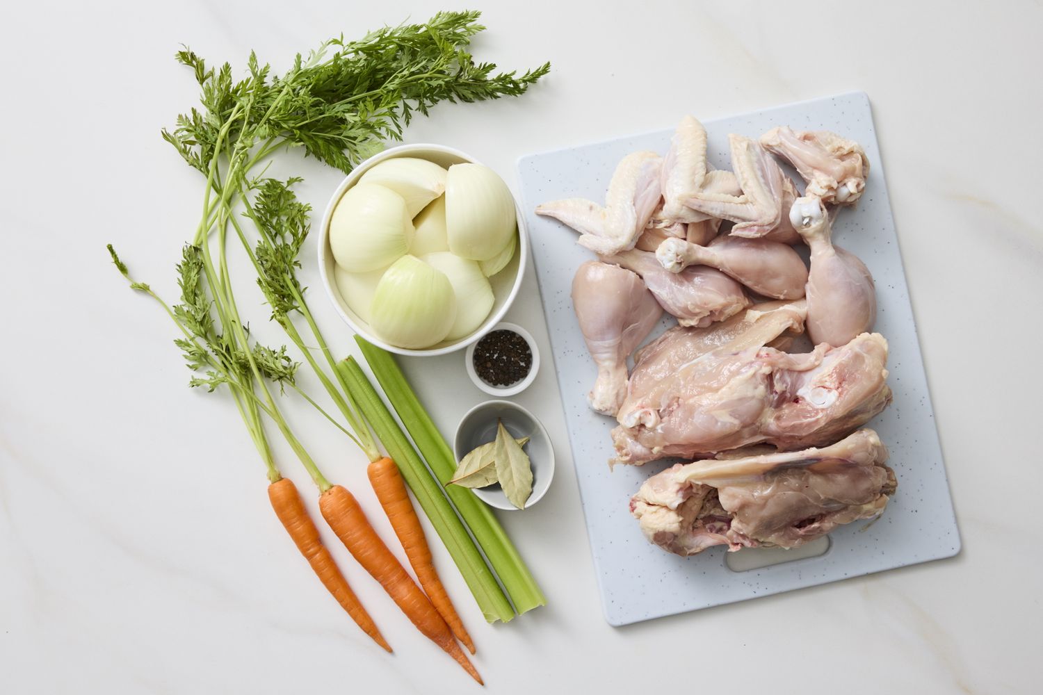 Ingredients for making chicken stock including raw chicken pieces carrots celery onions garlic bay leaves and black peppercorns