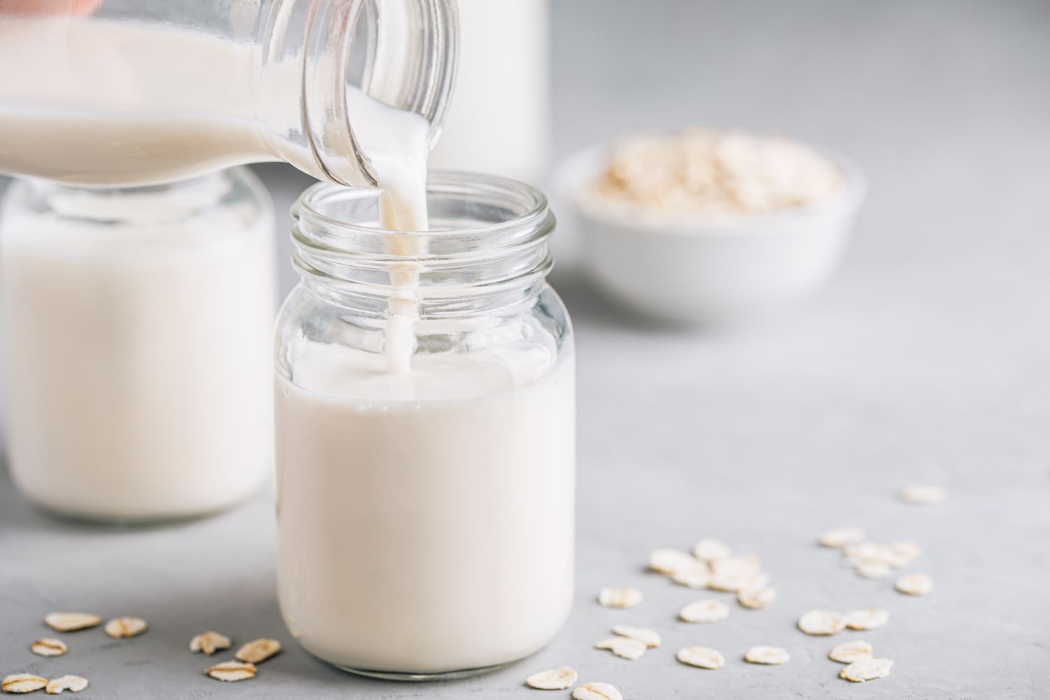Oat milk being poured into a glass jar with oats scattered around on a light surface