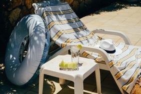 Lounge chair with a striped towel beside a table holding a drink and lime wedges and an inflatable tube nearby in a sunny outdoor setting