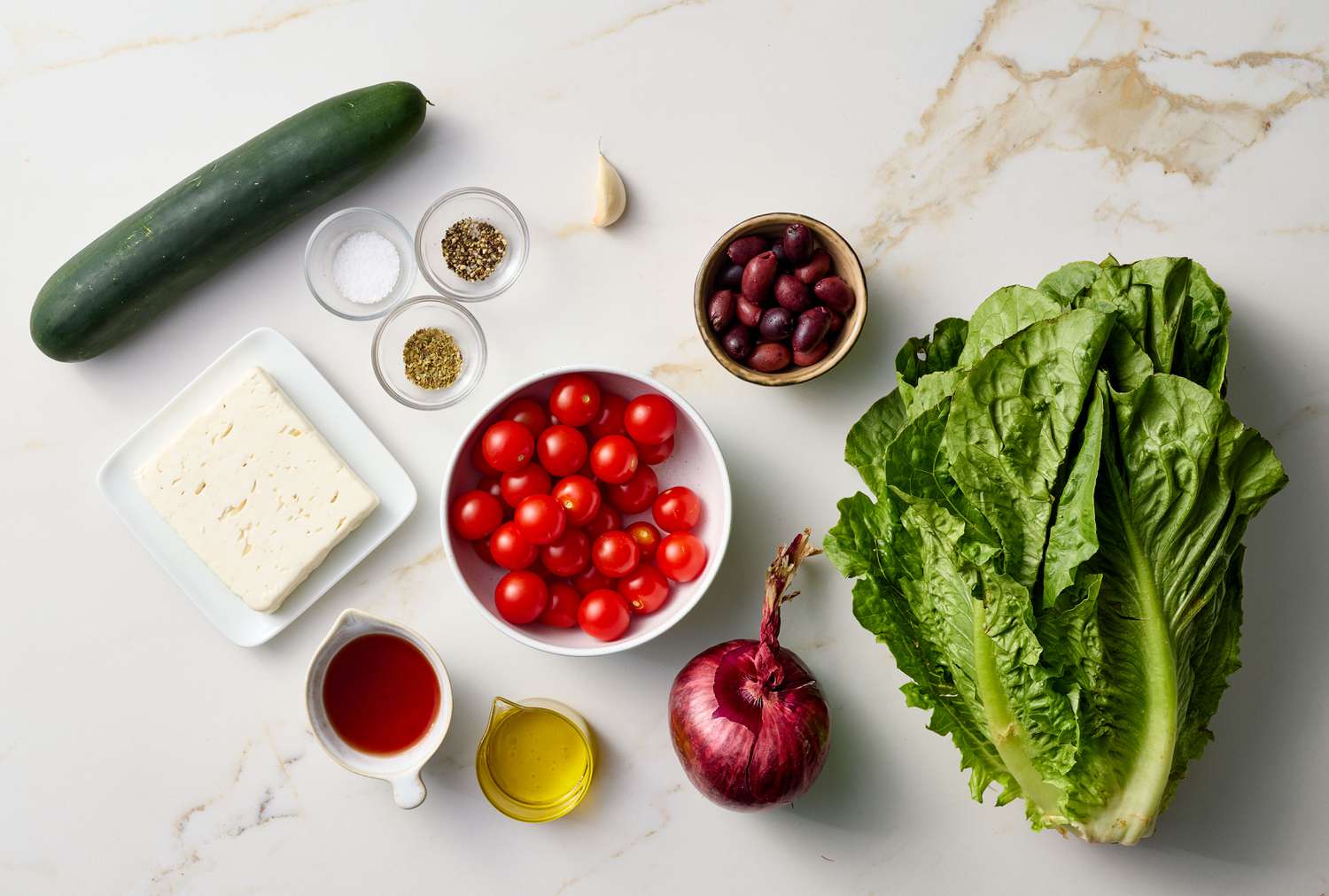 Ingredients for making Greek salad arranged on a white surface, including cucumbers, cherry tomatoes, olives, feta cheese, lettuce, and seasonings