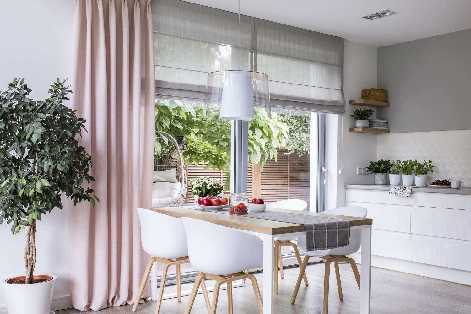 Gray roman shades and a pink curtain on big, glass windows in a modern kitchen and dining room interior with a wooden table and white chairs