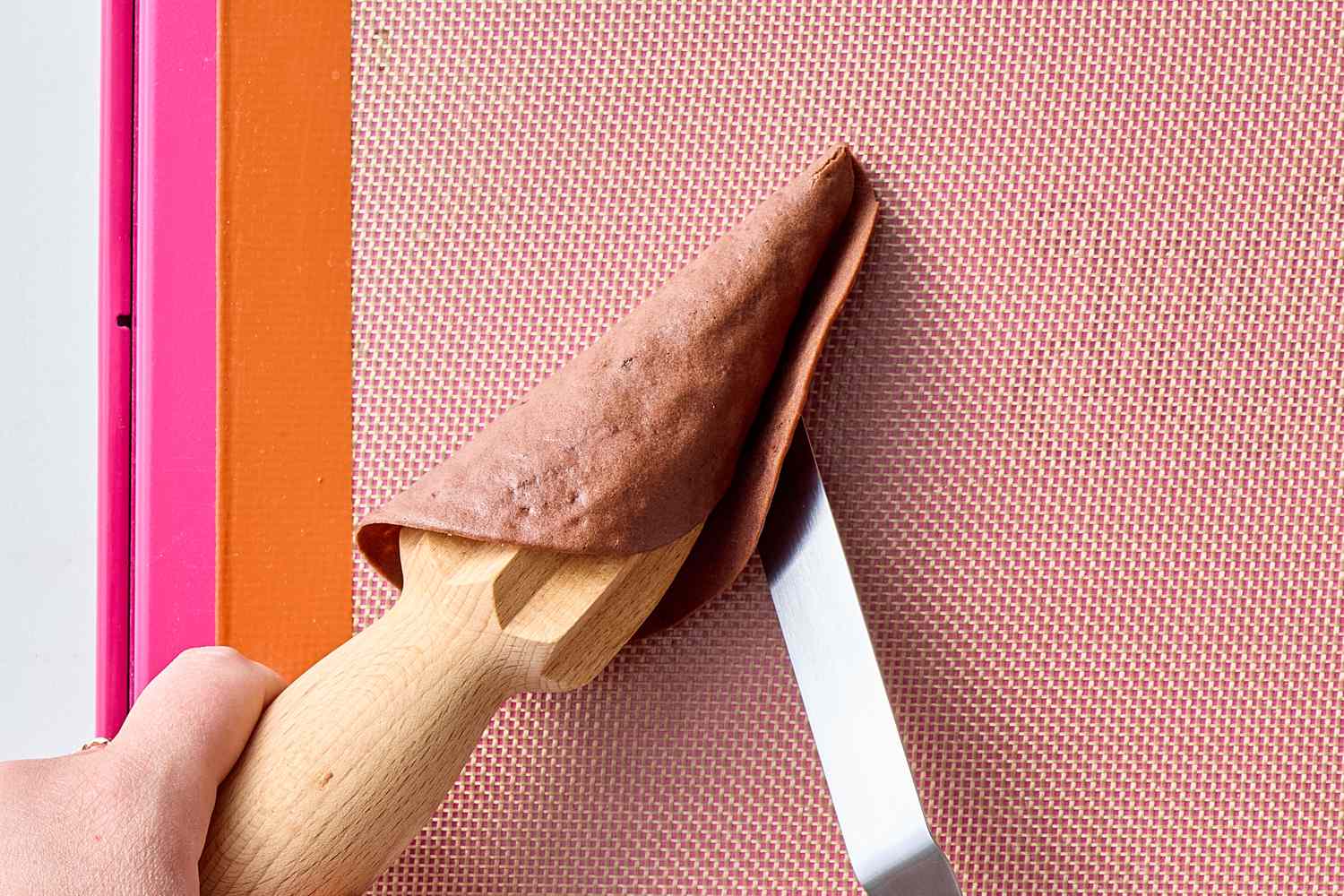 A hand shaping an ice cream cone out of a baked sheet using a wooden cone tool and a spatula on a silicone mat