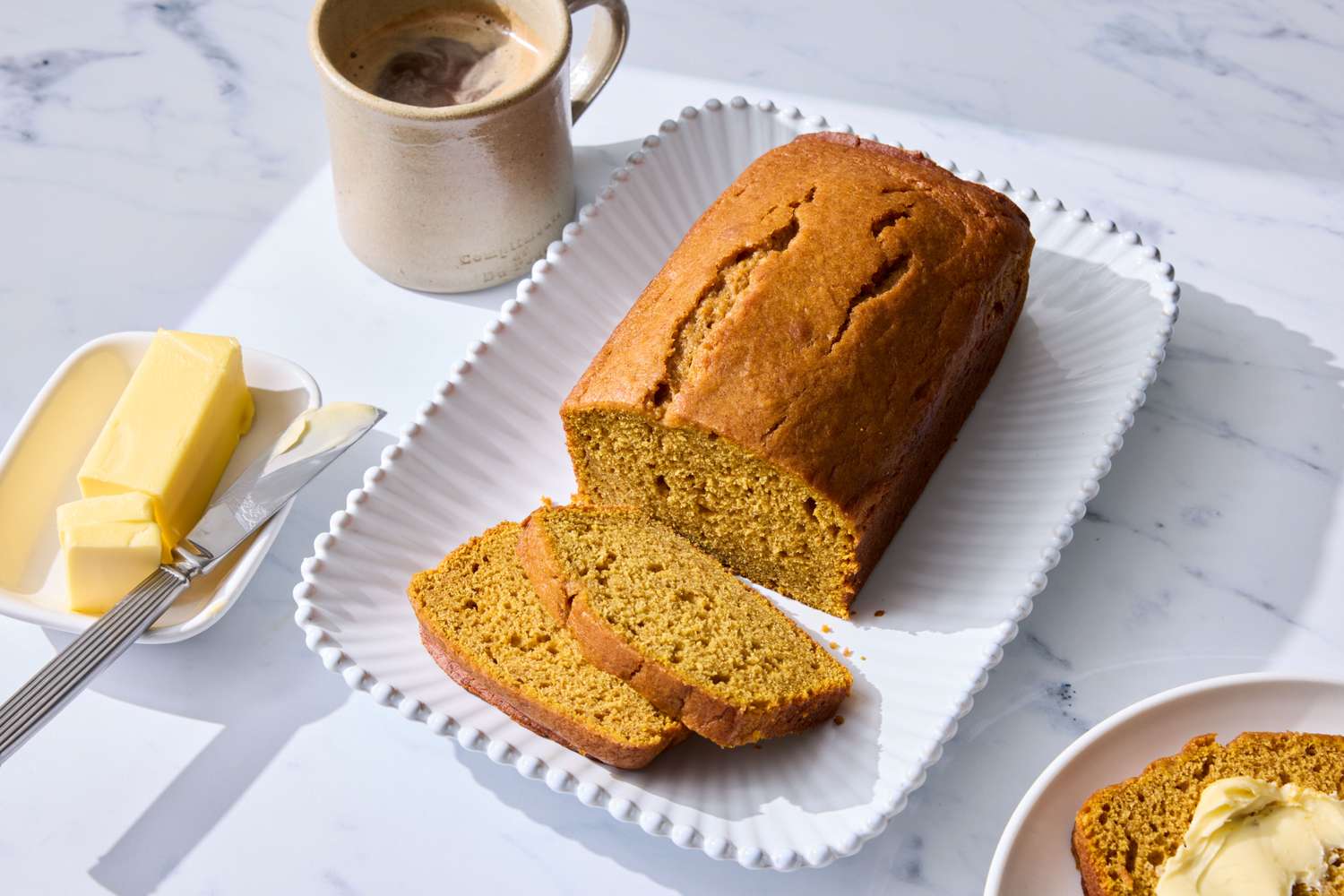 A loaf of pumpkin bread on a platter with a few slices cut accompanied by butter and a mug of coffee