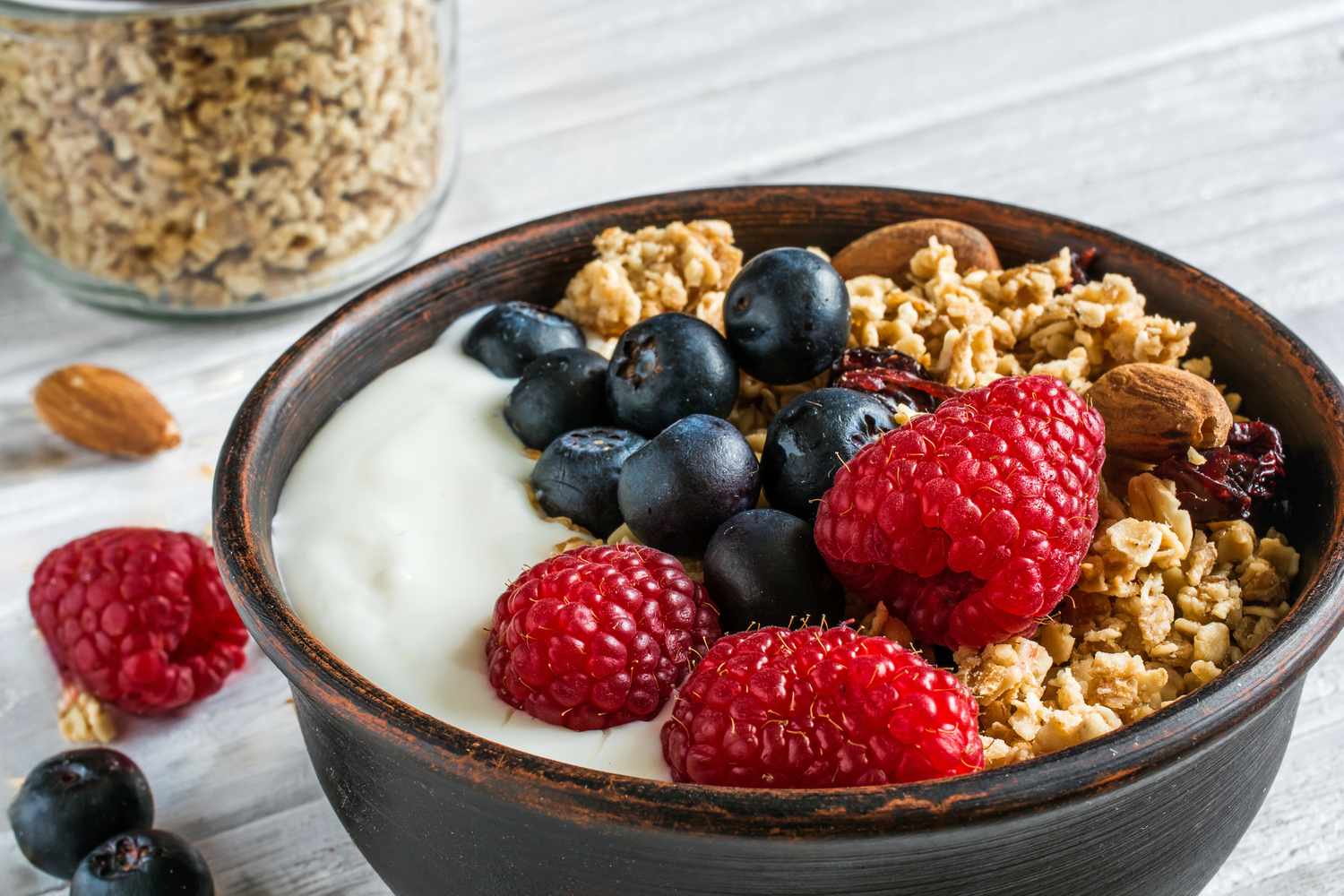 granola with greek yogurt, nuts and fresh berries in a bowl with cereal in jar over white wooden background