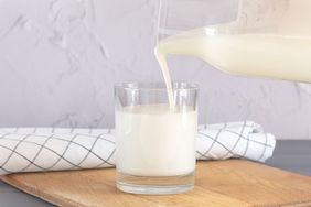 A glass of milk on a wooden surface being poured from a pitcher