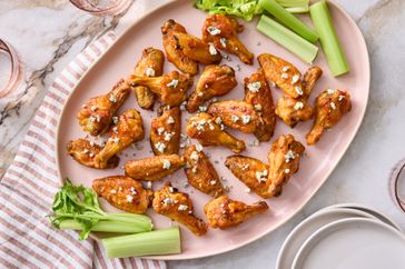 A platter of baked chicken wings topped with crumbled cheese accompanied by celery sticks on a pink plate with a striped cloth beneath