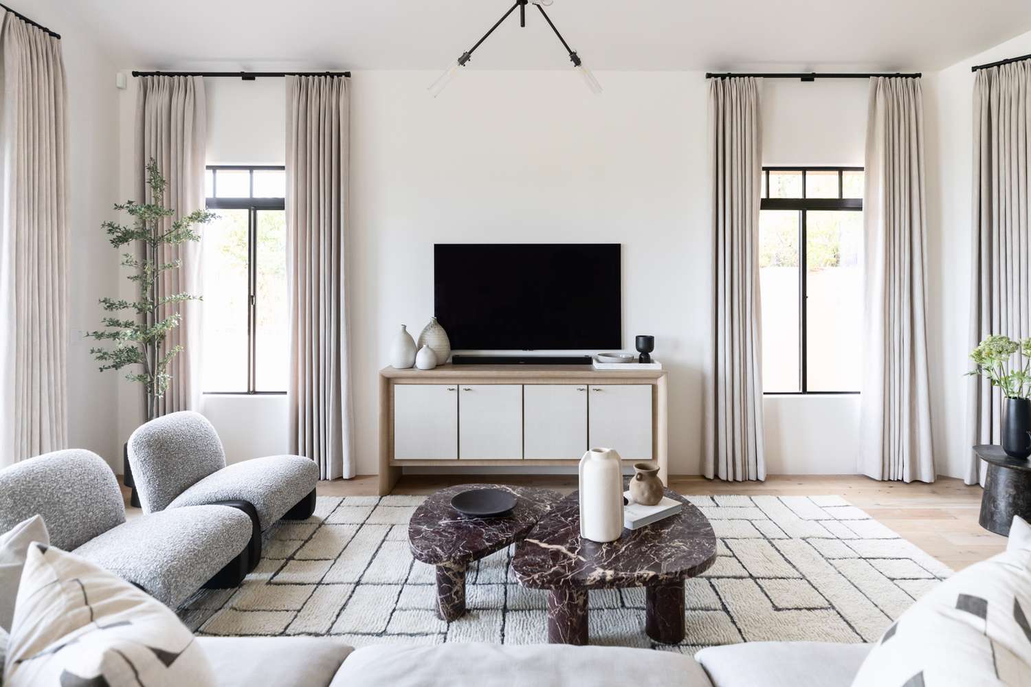Minimalist living room by Living with Lolo: gray bouclé chaise lounges, marbled coffee tables with organic shapes, beige curtains on large windows, potted greenery, geometric white rug, TV on white media console, and natural light create a calm, stylish space.