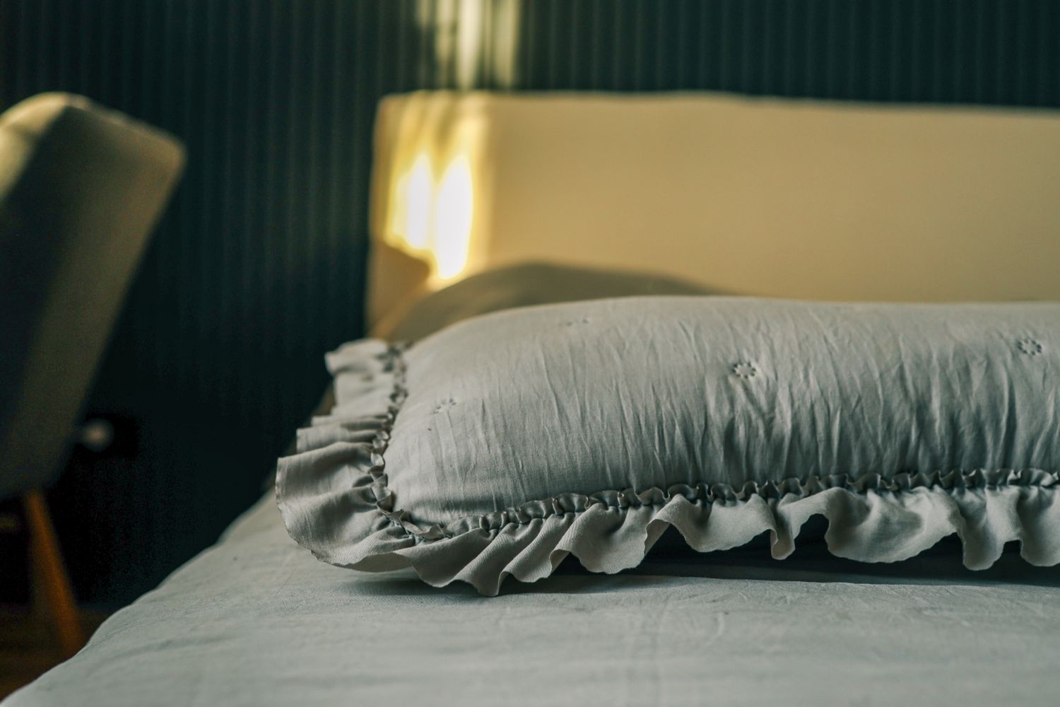 A bed with a decorative pillow in focus a chair and textured wall in the background