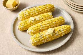 Four ears of corn on a plate topped with butter and ground pepper with a butter dish in the background