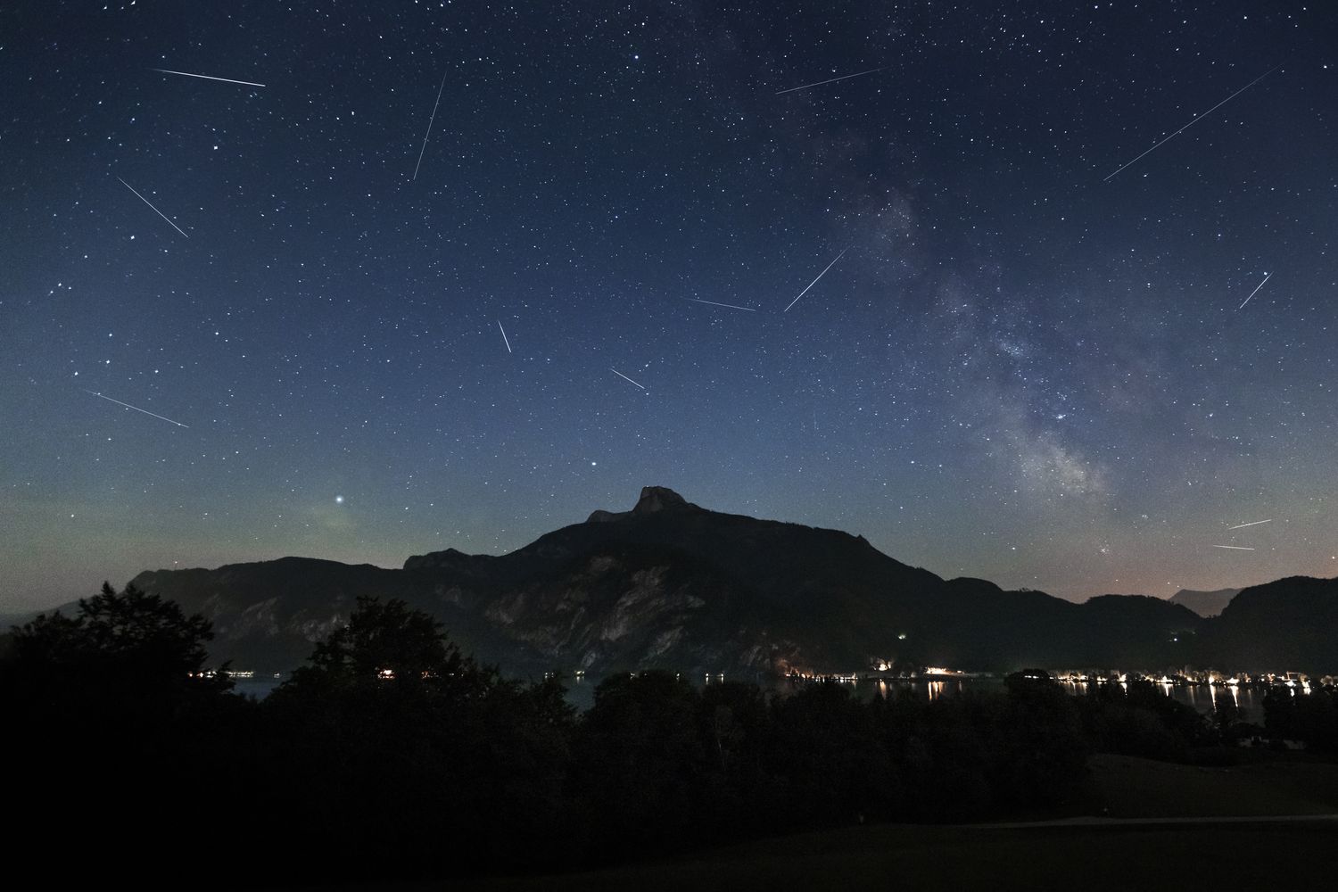 Night sky with a meteor shower silhouetted mountains with distant lights below