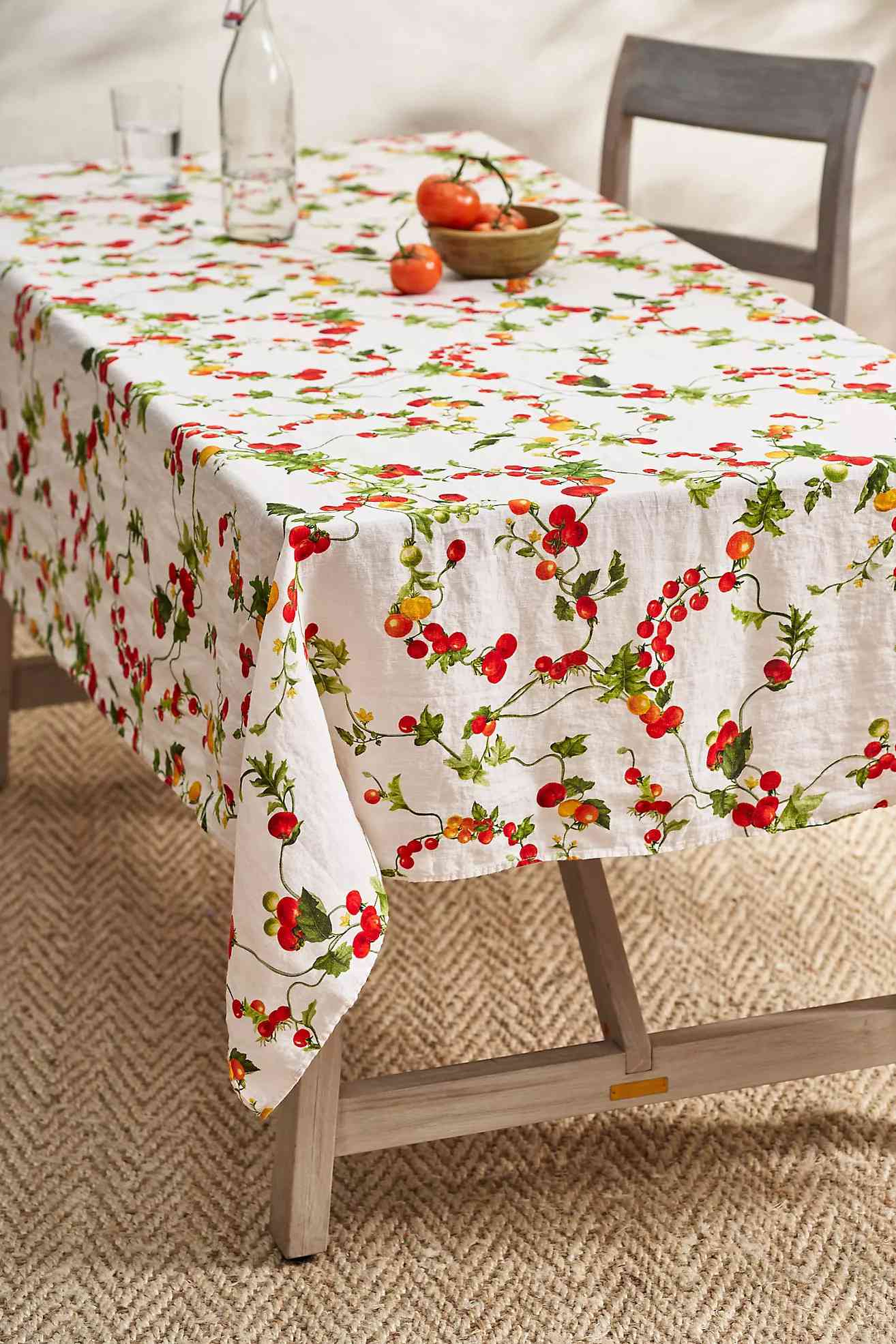 Table with a tablecloth decorated with a cherry pattern, a wooden chair, a glass bottle, a bowl, and tomatoes