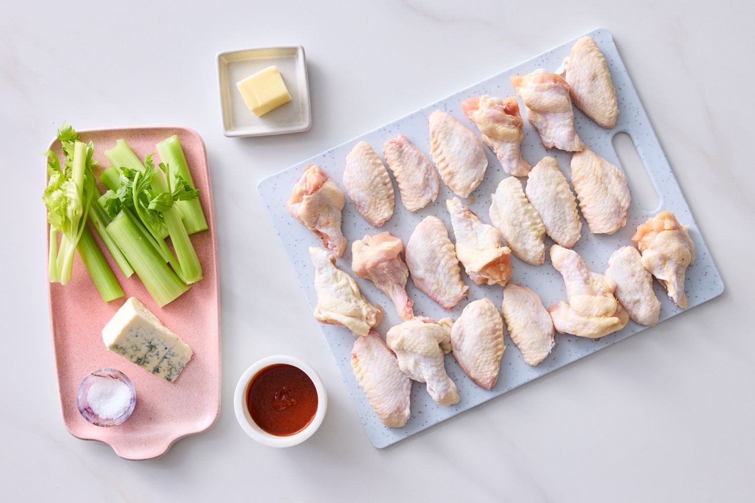 Raw chicken wings on a board with celery buffalo sauce blue cheese butter and salt arranged nearby