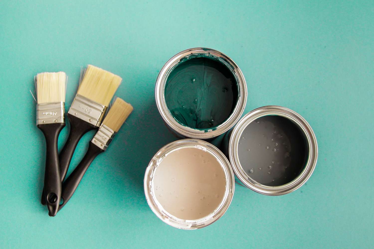Three paintbrushes next to three open cans of paint on a solid background