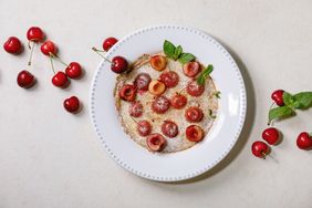 A plate with a pancake topped with cherries and garnished with mint surrounded by whole cherries and mint leaves