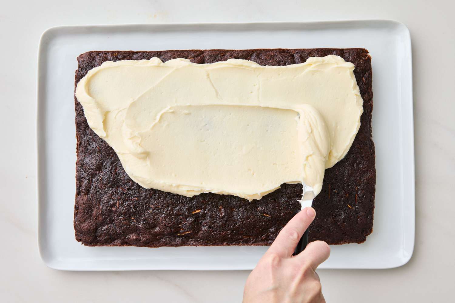 A hand spreading cream cheese frosting over a chocolate zucchini cake using an offset spatula
