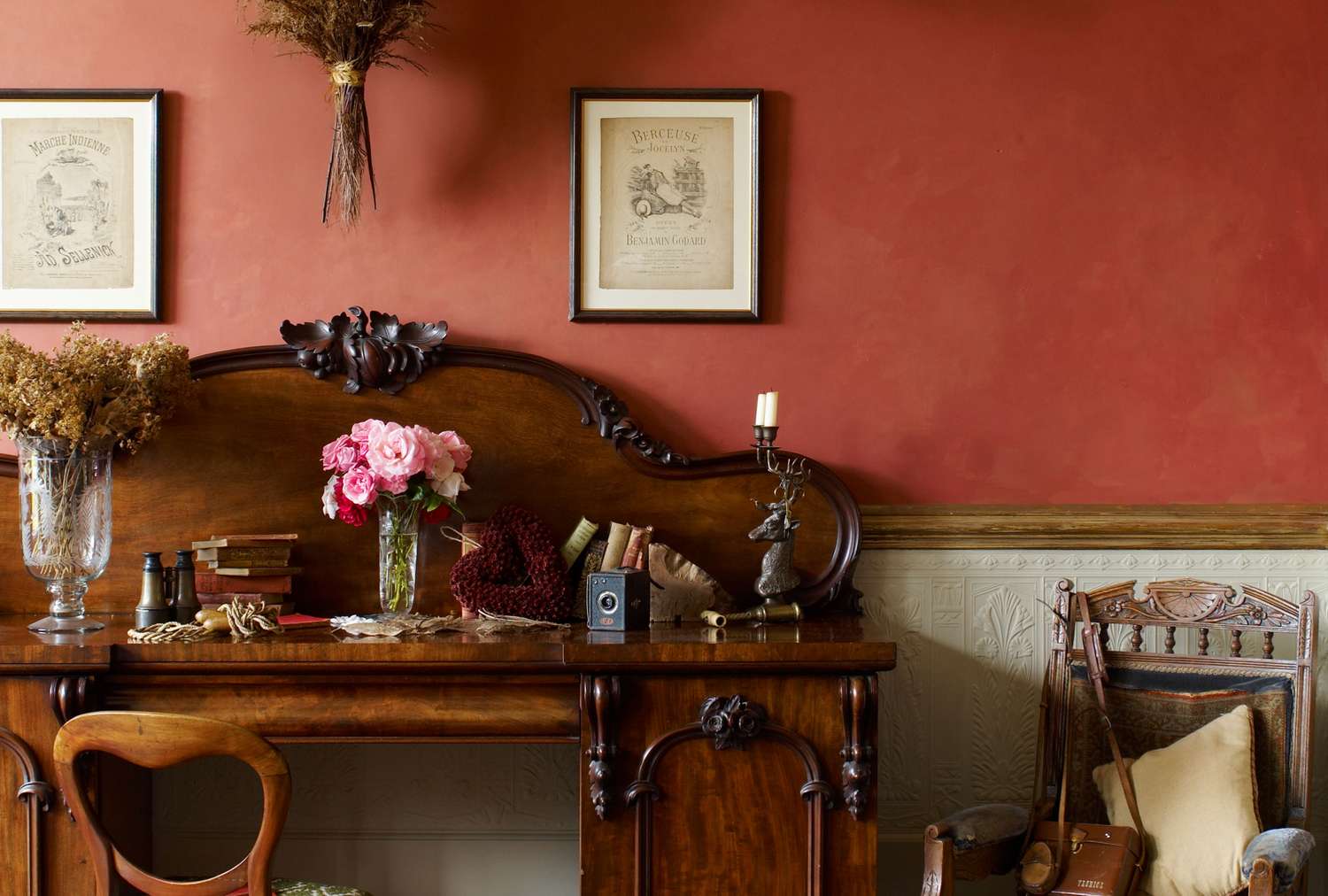 An ornate wooden desk with books flowers and a candleholder backed by a red wall with framed pictures and a decorative chair nearby