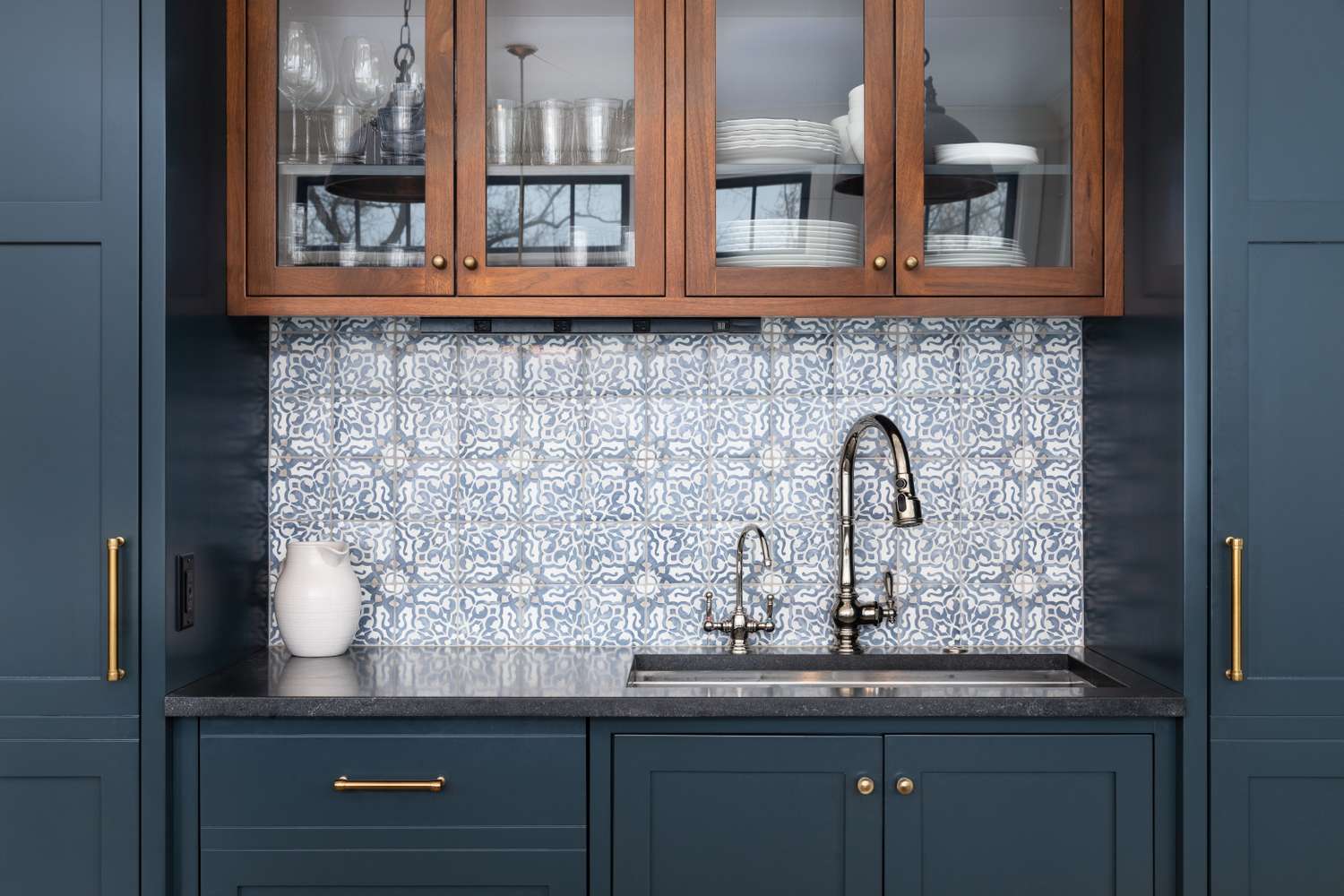 Kitchen interior with blue cabinets a patterned tile backsplash and a metal faucet over a sink featuring upper glassfront cabinets