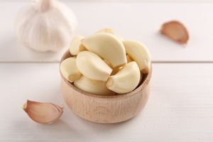 A wooden bowl filled with peeled garlic cloves placed on a surface with garlic bulbs and cloves around it