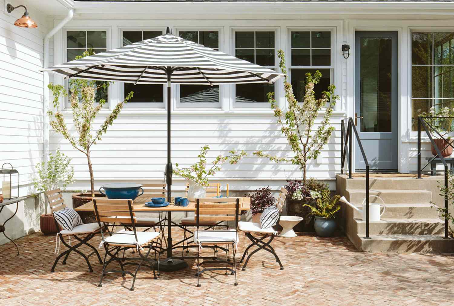 Outdoor patio area with a table chairs and an umbrella against a white house with lush plants and windows in the background
