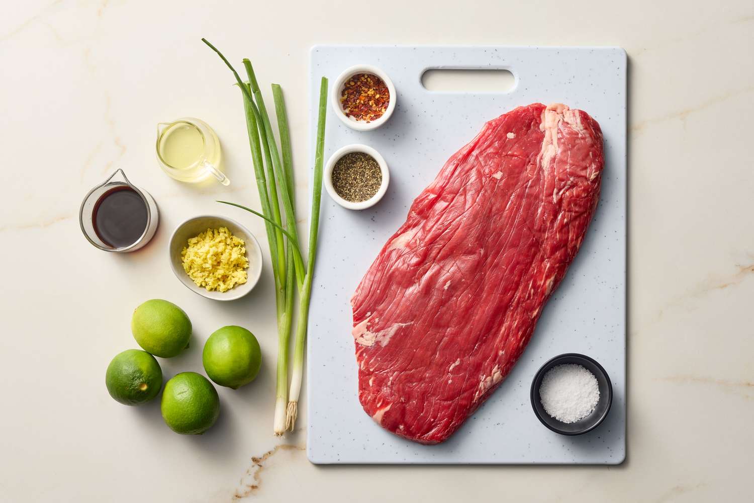 Flank steak on a cutting board with ingredients including lime, spring onion, spices, and oils arranged for a lime marinade preparation