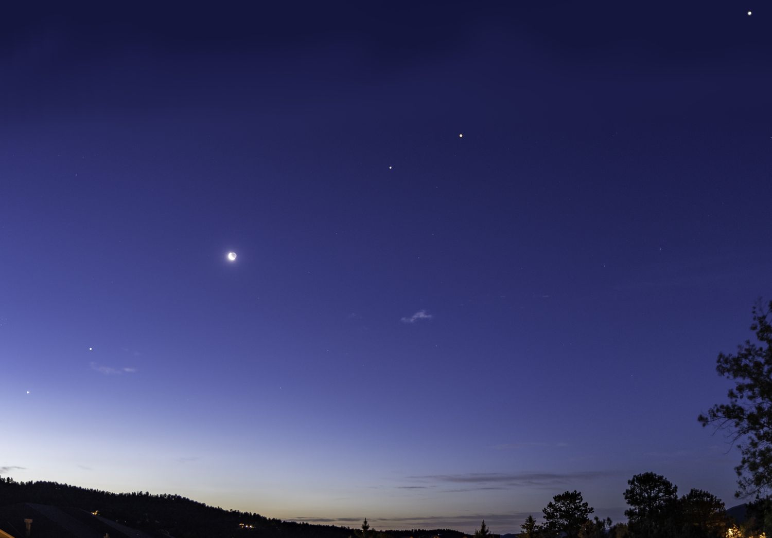 Night sky with celestial bodies and a crescent moon visible above a silhouette of treetops and hills