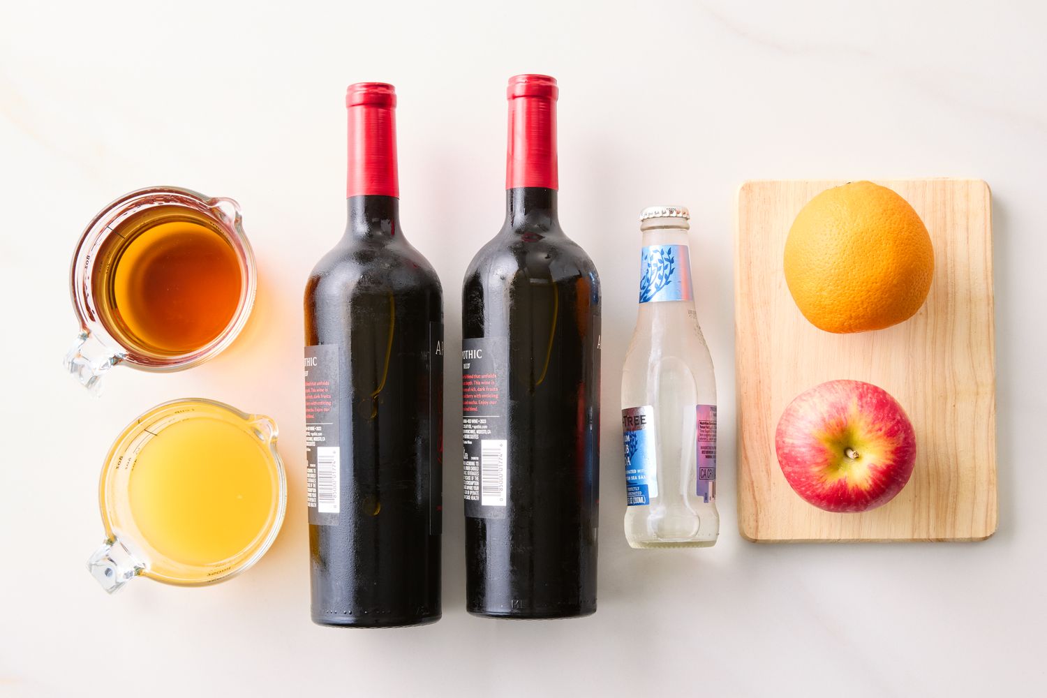 Ingredients for making red sangria including wine bottles fruit juices soda an orange and an apple on a cutting board
