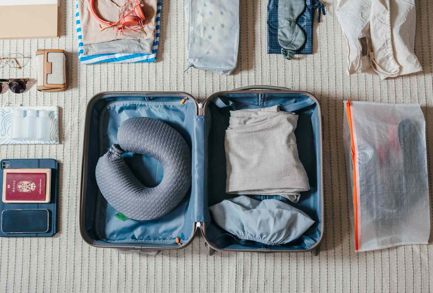 An organized suitcase with clothing a travel pillow and various items arranged neatly beside it on a carpeted surface