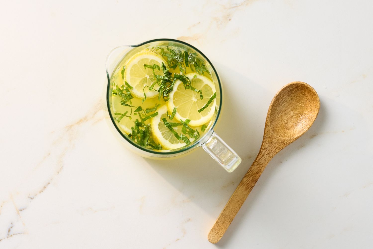 A pitcher of lemonade garnished with mint and lemon slices with a wooden spoon on a marble surface