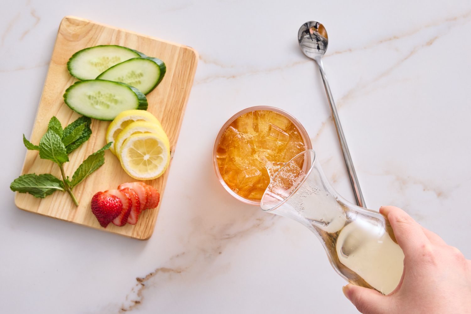 A glass with a beverage being poured a wooden board with sliced fruit and herbs and a mixing spoon on a counter