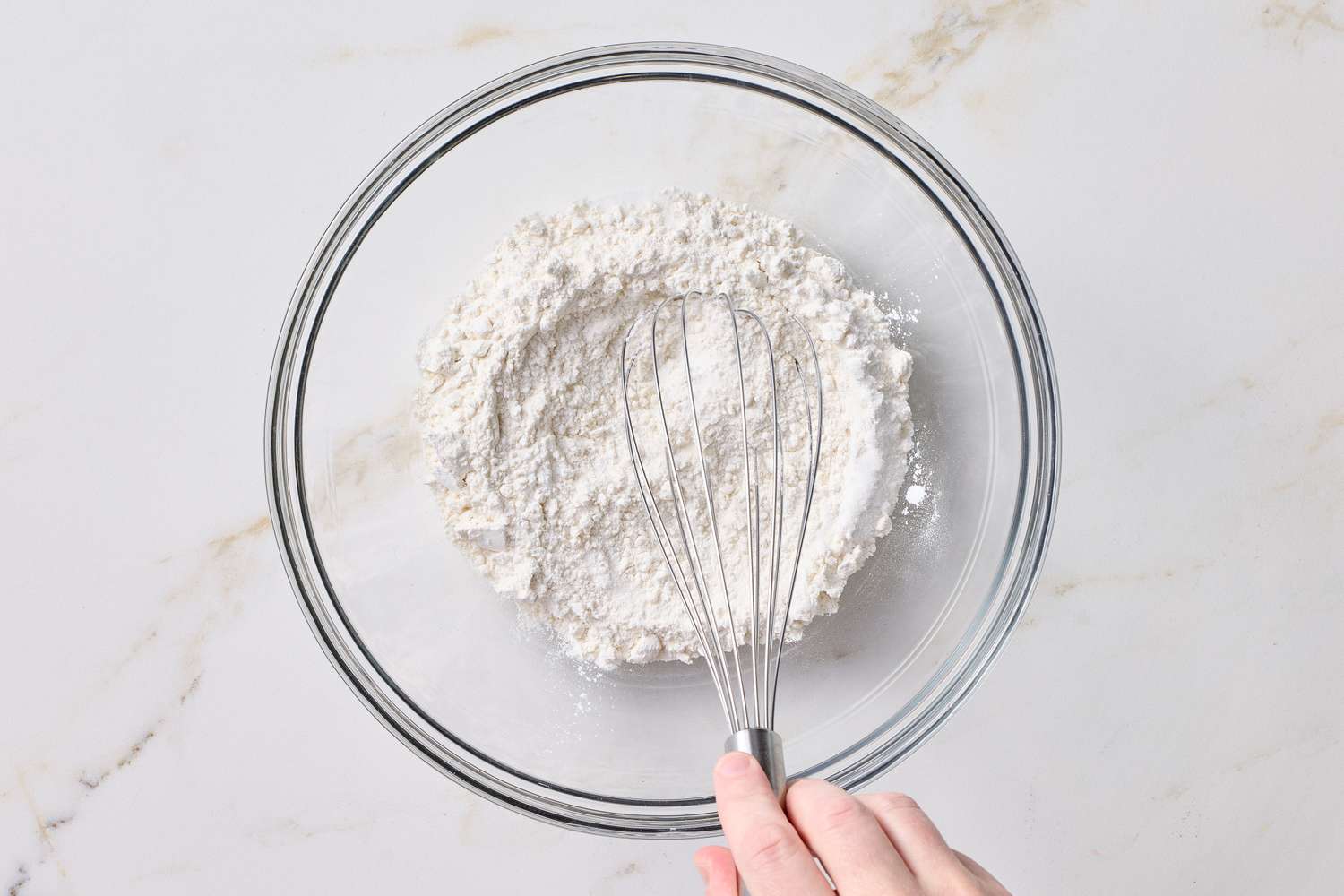 A hand holding a whisk in a glass bowl of flour mixture on a marble surface